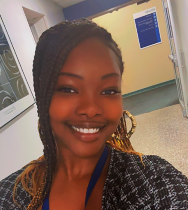 A close-up selfie of a person standing in a hallway indoors. The person is smiling at the camera and wearing a patterned black-and-white top and a blue lanyard with an ID badge. The hairstyle features long braids pulled over one shoulder. The background shows a light-colored hallway with a door and a wall-mounted sign visible behind the person.