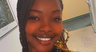A close-up selfie of a person standing in a hallway indoors. The person is smiling at the camera and wearing a patterned black-and-white top and a blue lanyard with an ID badge. The hairstyle features long braids pulled over one shoulder. The background shows a light-colored hallway with a door and a wall-mounted sign visible behind the person.