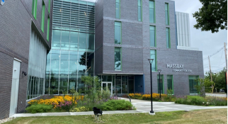 Modern MassBay Community College building with gray brick facade, green-trimmed vertical windows, a glass entryway, and landscaped grounds featuring walkways, grass, and yellow and purple flowers.